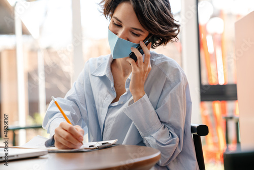 Happy girl in protective mask talking on cellphone while writing down notes