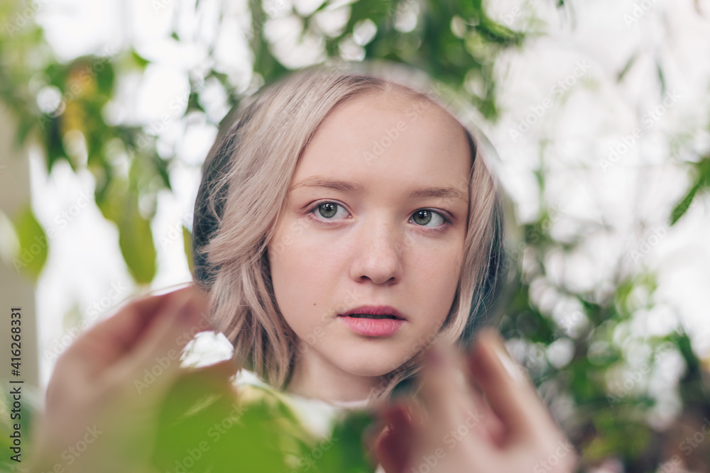 The reflection in the mirror of a beautiful blonde girl looking at herself. Plant leaves in the background. Psychological concept. Urban Jungle concept. The reflection in the mirror of a beautiful blonde girl looking at herself. Plant leaves in the background. Psychological concept. Urban Jungle concept.
