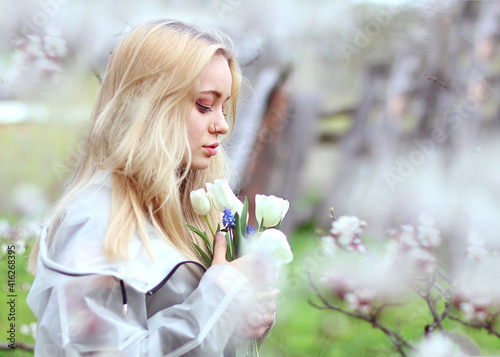A girl with white hair  in a transparent jacket  stands near a blossoming tree with white tulips. Spring mood