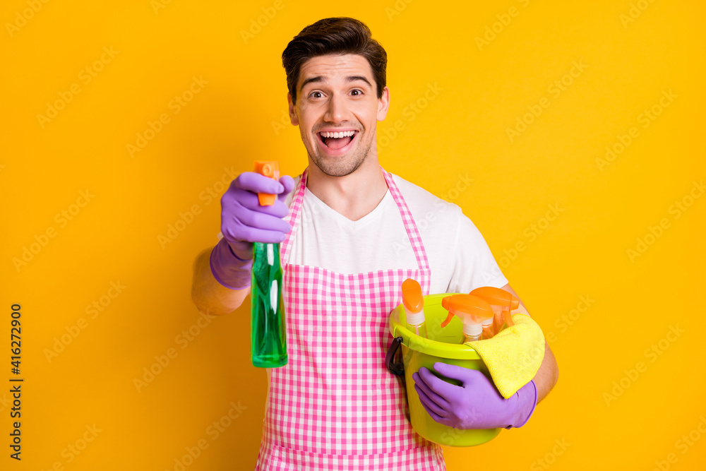 Photo of young happy excited smiling man in gloves hold cleaning splay and bucket with detergent isolated on yellow color background Photo of young happy excited smiling man in gloves hold cleaning splay and bucket with detergent isolated on yellow color background
