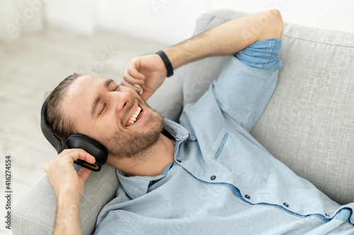 Attractive man with light brown hair and beard, singing his favorite song, smiling, listening music with headphones lying on the grey cozy couch, keeping his arms near the head, joyful, beautiful day