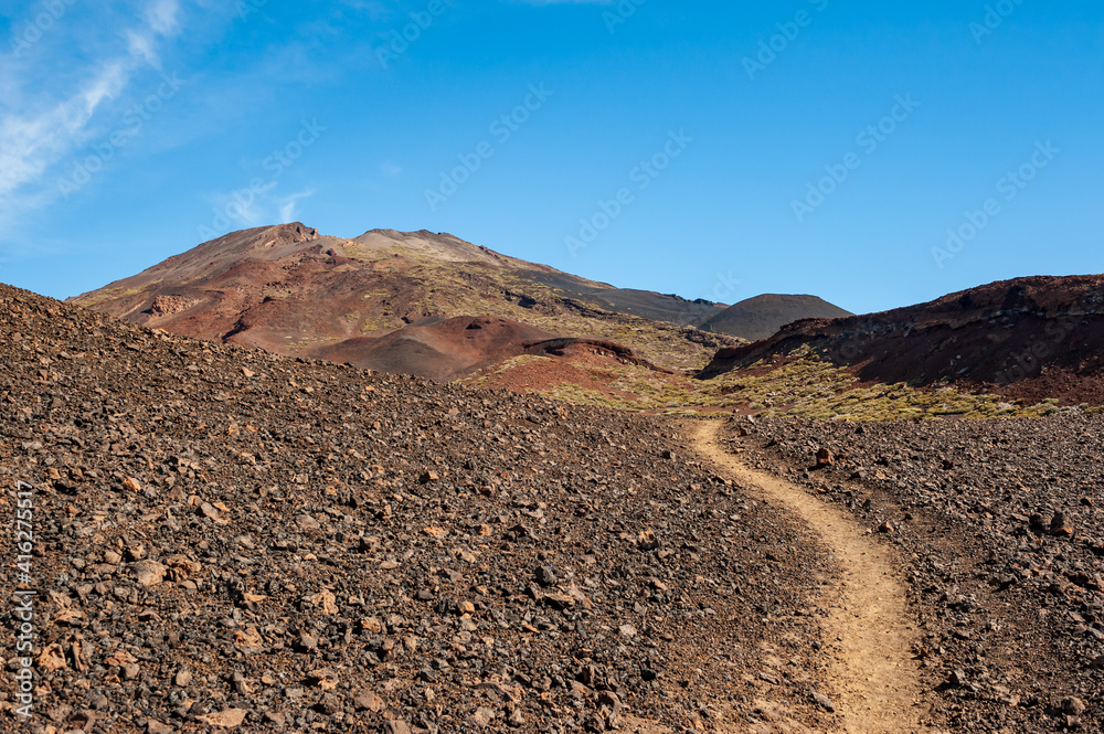 Path in desert and silhouette of volcano del Teide against  blue sky and female hiker with backpack. Pico del Teide mountain in El Teide National park. Tenerife, Canary Islands, Spain