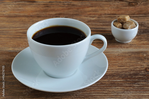 White porcelain cup and saucer with black coffee and cane brown sugar on wooden table