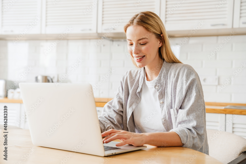Joyful blonde girl smiling and using laptop while sitting at kitchen Joyful blonde girl smiling and using laptop while sitting at kitchen