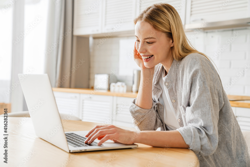 Joyful blonde girl smiling and using laptop while sitting at kitchen Joyful blonde girl smiling and using laptop while sitting at kitchen