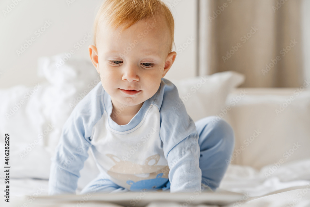 Focused blonde baby boy looking at tablet computer while playing on bed Focused blonde baby boy looking at tablet computer while playing on bed