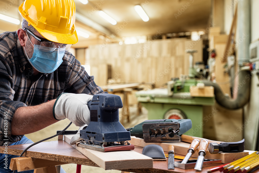 Carpenter worker at work in the carpentry workshop, wears helmet, goggles, leather gloves and surgical mask to prevent coronavirus infection. Preventing Pandemic Covid-19 at the workplace. Carpenter worker at work in the carpentry workshop, wears helmet, goggles, leather gloves and surgical mask to prevent coronavirus infection. Preventing Pandemic Covid-19 at the workplace.