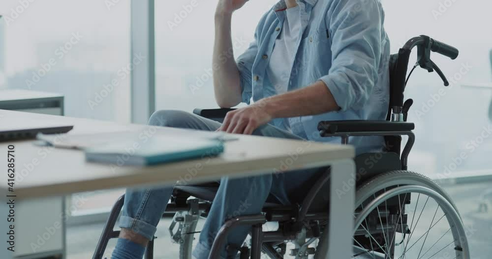Young man disabled in black wheelchair sitting in corporate office talking to friend mobile phone internet communicating calling indoors. Corporate people. Young man disabled in black wheelchair sitting in corporate office talking to friend mobile phone internet communicating calling indoors. Corporate people.