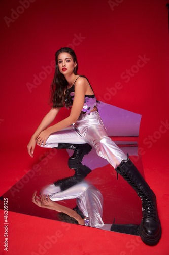 Fashionable woman in silver jeans, sequins top and black boots. Girl with make-up and long wet brunette hair sitting and posing at flexible mirror and red background