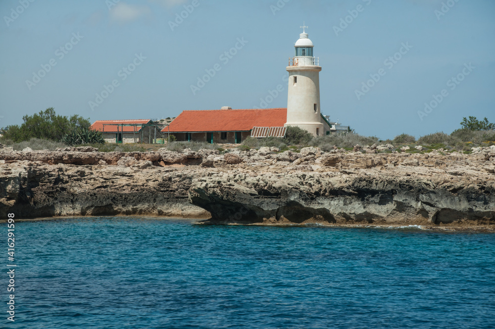View of the small white lighthouse on the rock from sea . Landscape with turquoise sea and grottoes on coast of Cyprus