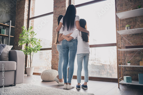 Full size rear portrait of happy lady little daughters standing near window hugging spend vacation home indoors