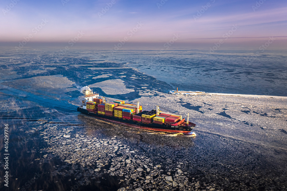 Cargo ship driving through frozen sea with colorful containers. Dark waters with ice blocks floating in sea.