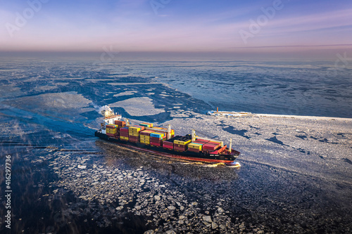 Obraz na płótnie Cargo ship driving through frozen sea with colorful containers. Dark waters with ice blocks floating in sea. w salonie