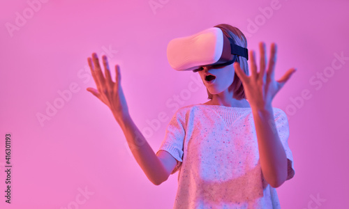 Attractive surprised shocked woman in casual wear using virtual reality glasses looks at her hands isolated on neon pink background. VR headset. Future technologies. © Valerii Apetroaiei