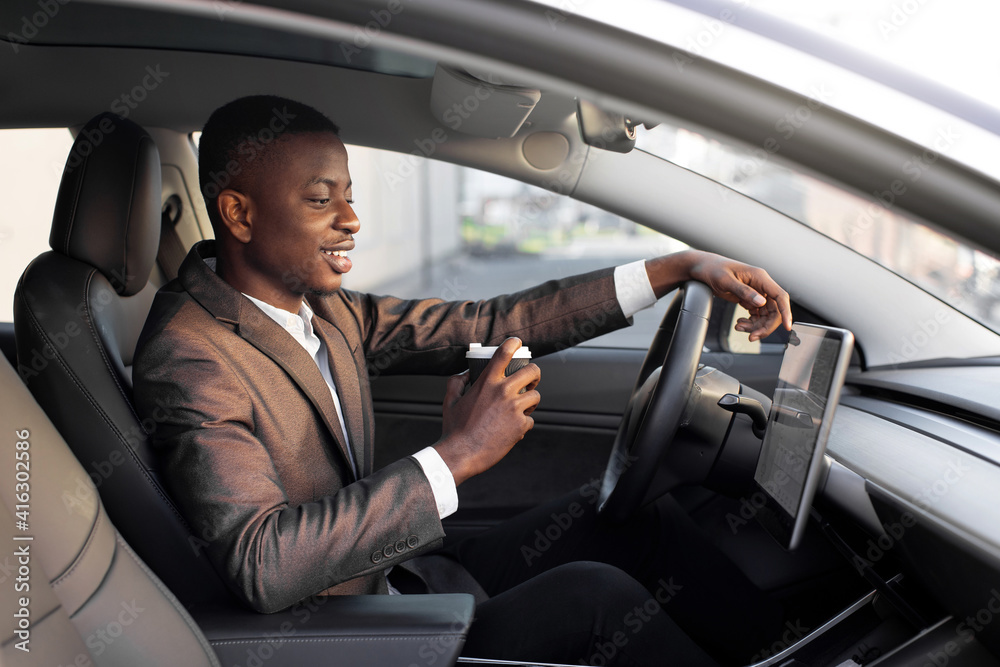 Young excited African businessman, having his lunch break to drink take away coffee, while sitting in modern electric car with self-steering system, and looking at car touchscreen display Young excited African businessman, having his lunch break to drink take away coffee, while sitting in modern electric car with self-steering system, and looking at car touchscreen display
