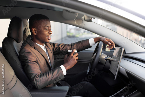 Young excited African businessman, having his lunch break to drink take away coffee, while sitting in modern electric car with self-steering system, and looking at car touchscreen display © sofiko14