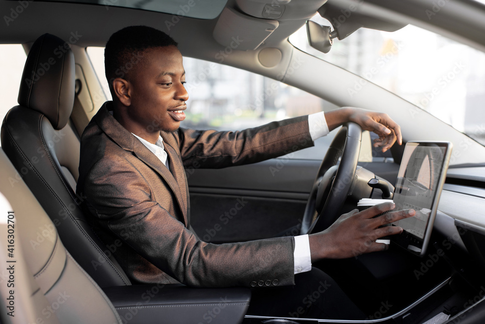 Smiling African American young businessman, dirnking coffee to go during break, sitting in the cabin of a comfortable modern electric car and touching the screen of navigation self-driving autopilot