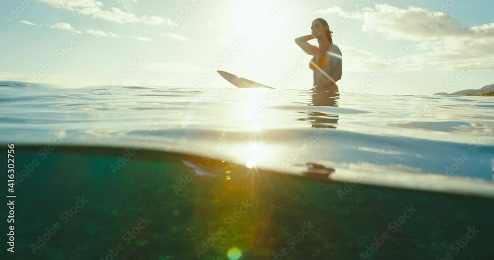 Beautiful female surfer sitting on surfboard in ocean at sunset waiting for the next wave. Slow motion shot on RED cinema camera.