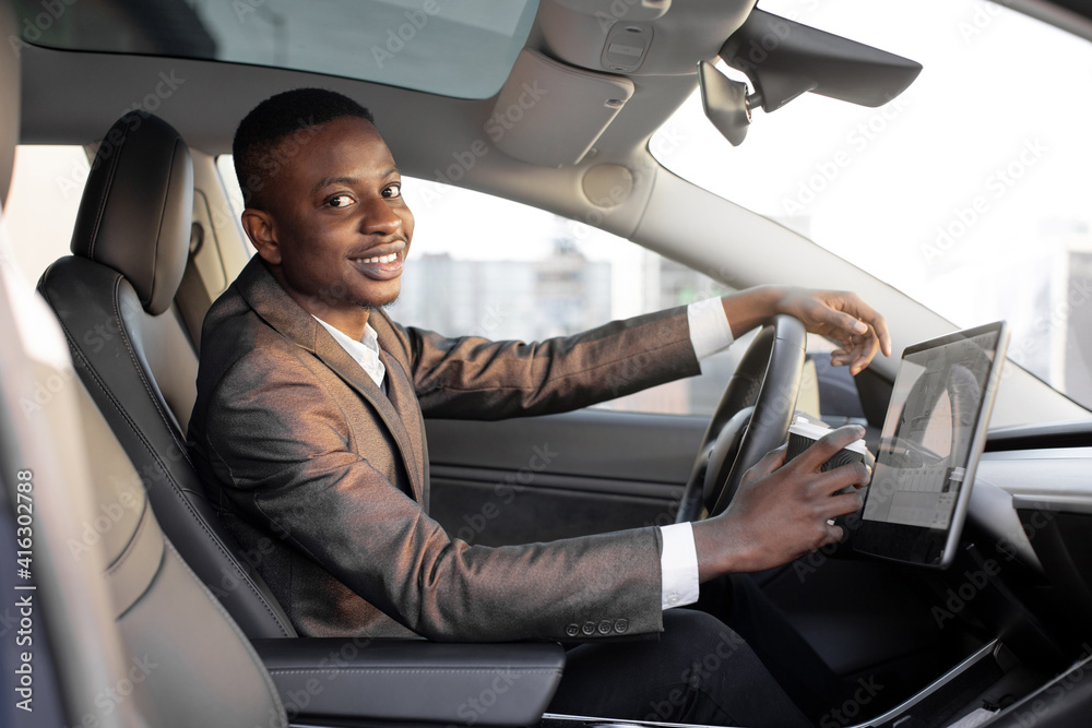 Happy smiling African American male driver sitting behind the self-driving steering wheel of an autonomous electric modern car. Happy guy holds coffee cup and smiles to camera in modern electric car Happy smiling African American male driver sitting behind the self-driving steering wheel of an autonomous electric modern car. Happy guy holds coffee cup and smiles to camera in modern electric car