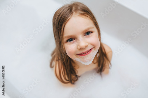 A small and beautiful red-haired girl, a child bathes, is washed in a white bath with soap foam. Happy childhood.