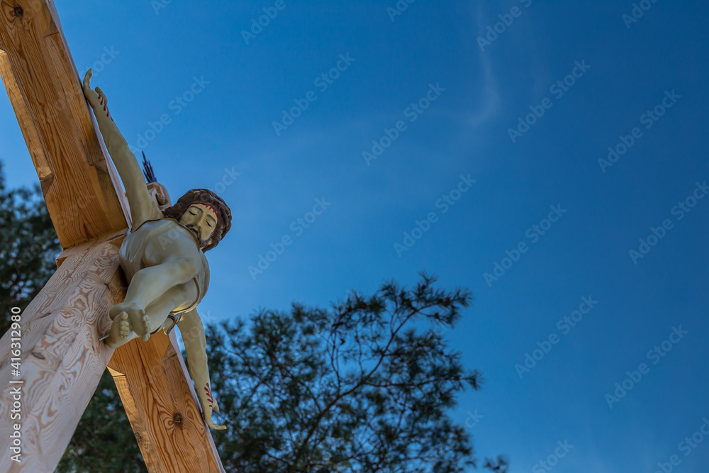 Fototapeta premium A figure of Jesus on a wooden roadside cross against a blue sky. Middle of the day, perfect lighting conditions.
