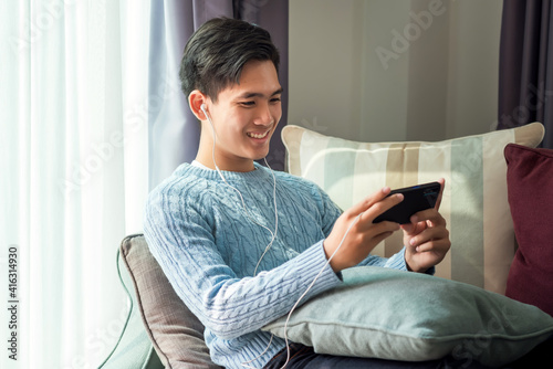Young Asian man lying on the sofa, wearing headphones, playing phone at home.