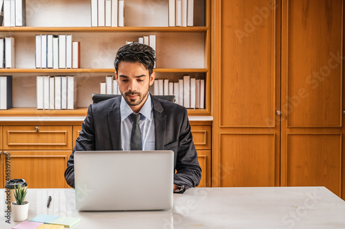 A businessman or lawyer in a suit sitting at his desk in an office looking at the laptop screen typing.