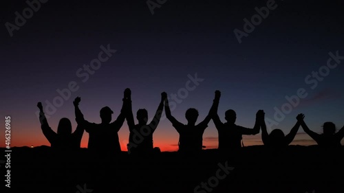 Silhouette showing group of people holding hands together as a team in front of colorful sunset. Close up pan shot.