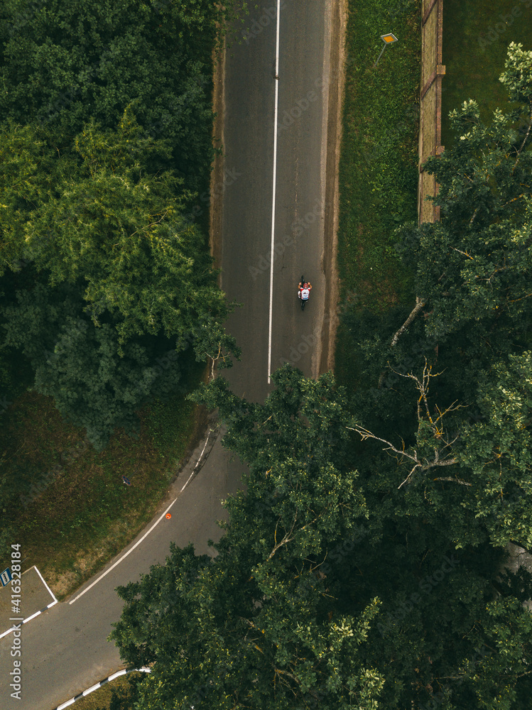 AERIAL, TOP DOWN: Flying above person riding their motorcycle along a curve of an empty asphalt road leading through the dark green forest.