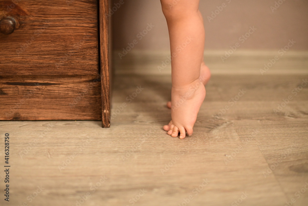 The baby's small feet are on the floor standing in tip toe in front of the dresser.