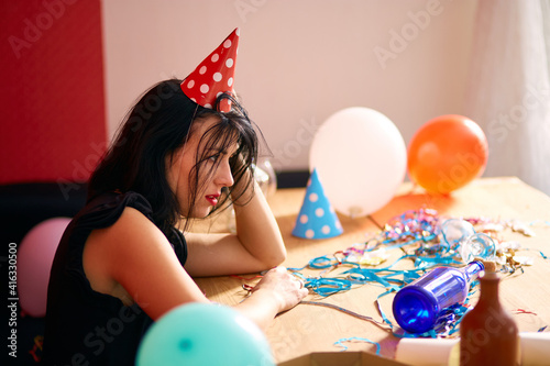 Young woman with red rubbed lipstick and cap, sitting tired at table in messy room after birthday party