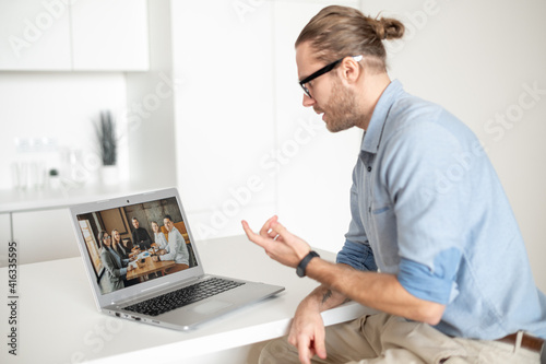Young diligent employee with glasses and hair pulled up, sitting at the laptop on a business meeting with a with potential clients and partners, looking at the laptop screen, working online
