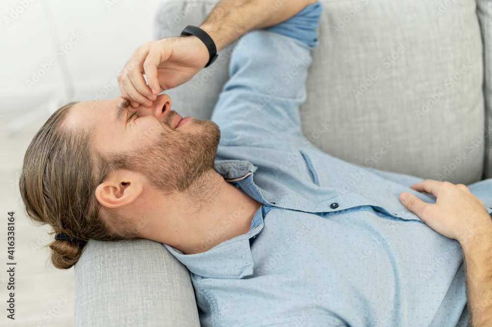 Fototapeta premium Attractive man with light brown hair pulled back, beard, lying on the grey cozy couch, with pleasure smile, keeping arm on the face, remembering this fantastic day, got long-awaited job, after date