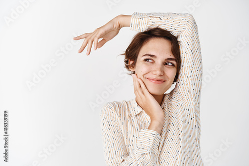Young happy woman in blouse, posing relaxed and sensual on white background, touching clean soft face and smiling, looking aside at empty space for logo, white background