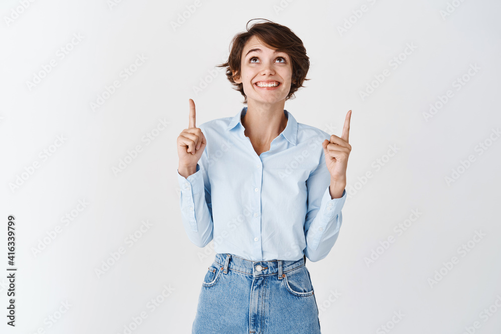 Excited lucky woman in blue collar shirt, pointing and looking up with cheerful smile, checking out company banner, standing on white background Excited lucky woman in blue collar shirt, pointing and looking up with cheerful smile, checking out company banner, standing on white background