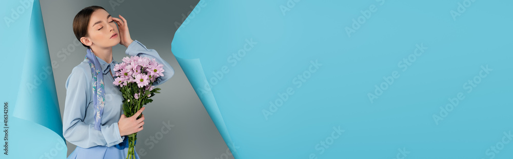 elegant woman posing with closed eyes while holding flowers near hole in blue paper on grey background, banner elegant woman posing with closed eyes while holding flowers near hole in blue paper on grey background, banner