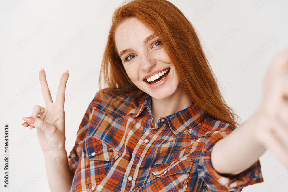 Beautiful redhead girl smiling happy and showing peace sign while taking selfie on smartphone, front camera view, white background Beautiful redhead girl smiling happy and showing peace sign while taking selfie on smartphone, front camera view, white background