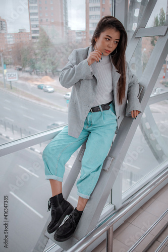 brunette with a Japanese appearance in a light sweater and a gray jacket and jeans against the backdrop of the city