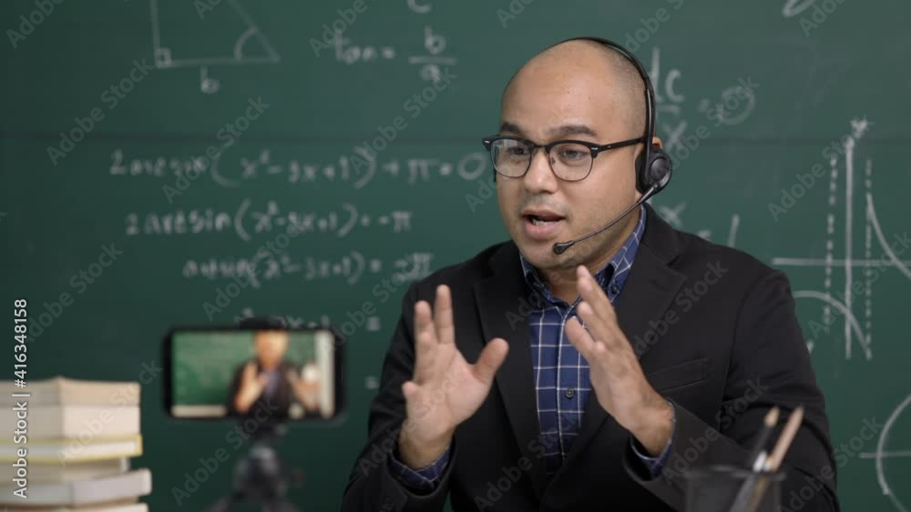 Indian young teacher man sitting wearing headset teaching online video conference live stream by smartphone. Asian teacher teaching mathematics class webinar online for students learning. Indian young teacher man sitting wearing headset teaching online video conference live stream by smartphone. Asian teacher teaching mathematics class webinar online for students learning.