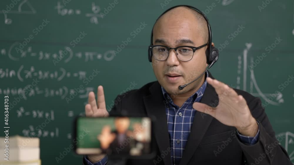 Indian young teacher man sitting wearing headset teaching online video conference live stream by smartphone. Asian teacher teaching mathematics class webinar online for students learning. Indian young teacher man sitting wearing headset teaching online video conference live stream by smartphone. Asian teacher teaching mathematics class webinar online for students learning.