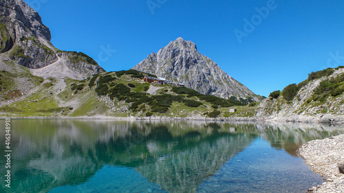 See mit Spiegelung in den Bergen und einer Berghütte bei blauem Himmel