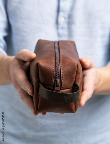 Fotografie Man's brown leather personal cosmetic bag or pouch for toiletry accessory in a men's hands in blue shirt