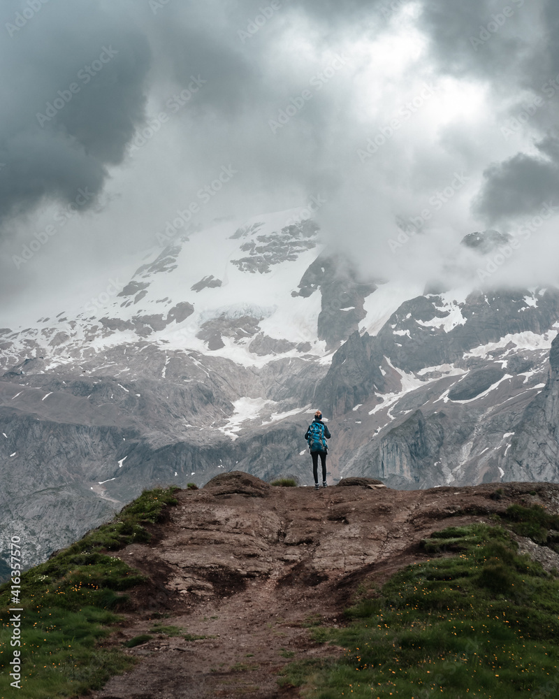 Hiking woman standing on a mountain ridge in front of marmolada glacier. Low clouds and a dramatic scenery. Dolomites, Alto Adige, Italy. 

