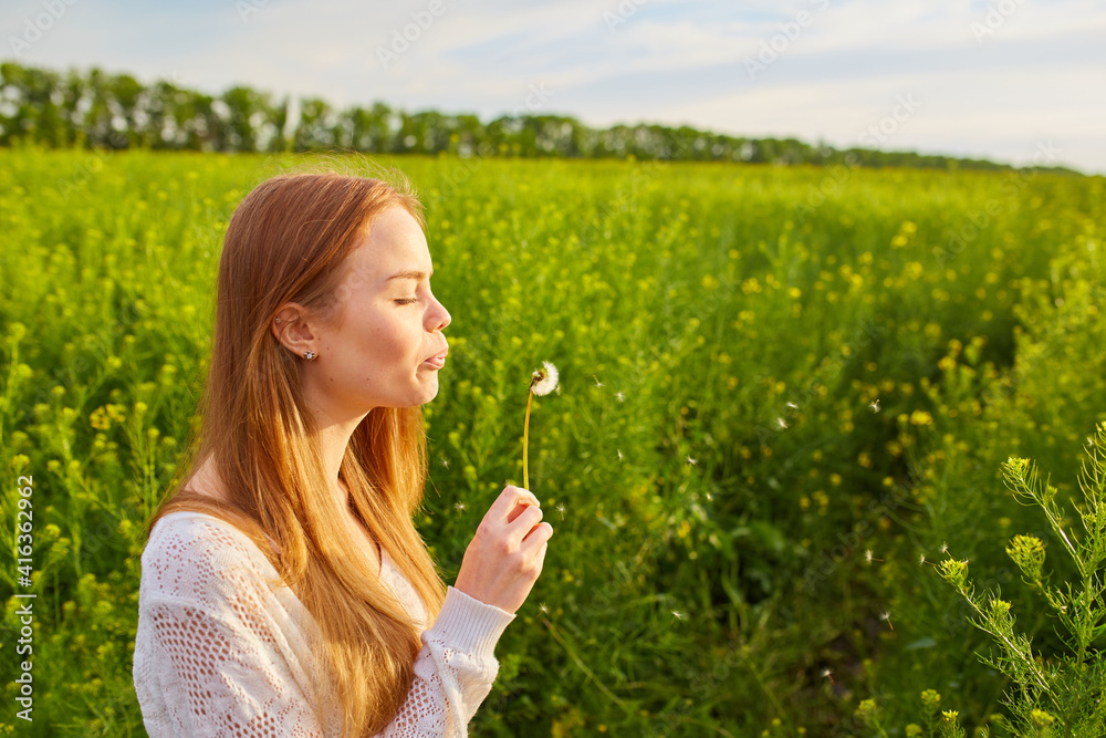 Young spring fashion redhead woman blowing dandelion in spring garden. Springtime, summertime. Trendy girl in summer landscape background. Allergic to pollen of flowers. Young spring fashion redhead woman blowing dandelion in spring garden. Springtime, summertime. Trendy girl in summer landscape background. Allergic to pollen of flowers.