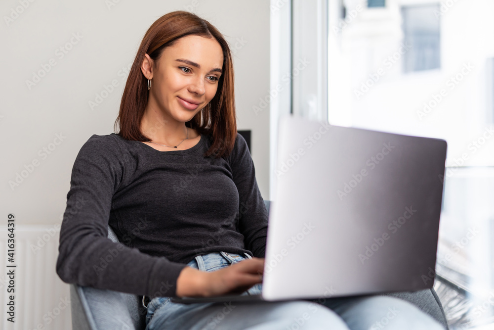Fototapeta premium Young smiling woman sitting on modern chair near the window in light cozy room at home working on laptop in relaxing atmosphere