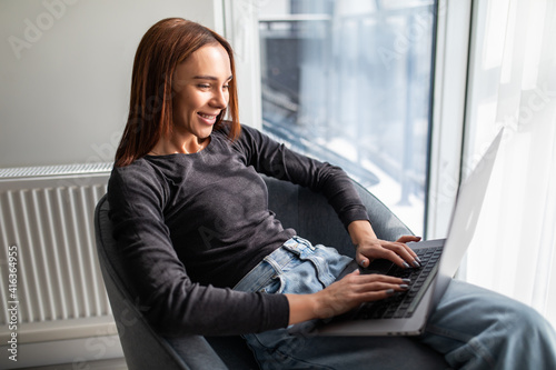 Young smiling woman is sitting on modern chair near the window in light cozy room at home. She is working on laptop in relaxing atmosphere