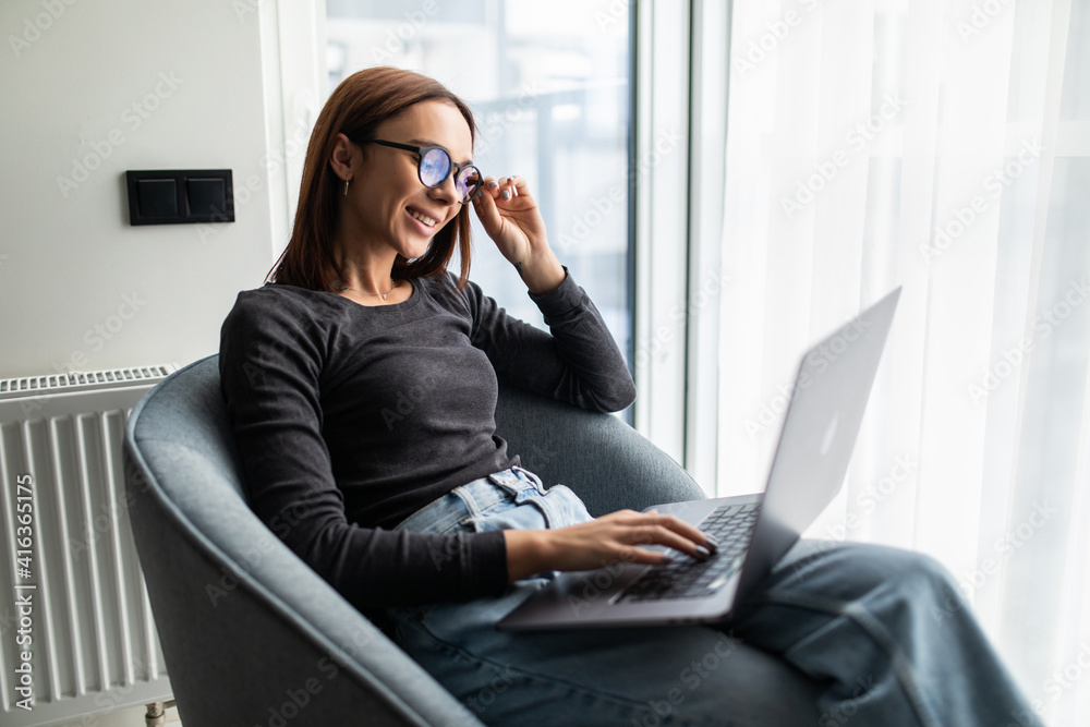 Young smiling woman sitting on modern chair near the window in light cozy room at home working on laptop in relaxing atmosphere Young smiling woman sitting on modern chair near the window in light cozy room at home working on laptop in relaxing atmosphere