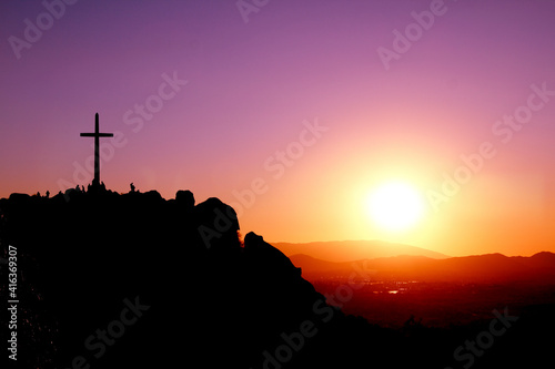 Cross on top of a mountain with silhouettes of people and a purple, orange, and yellow sunset.