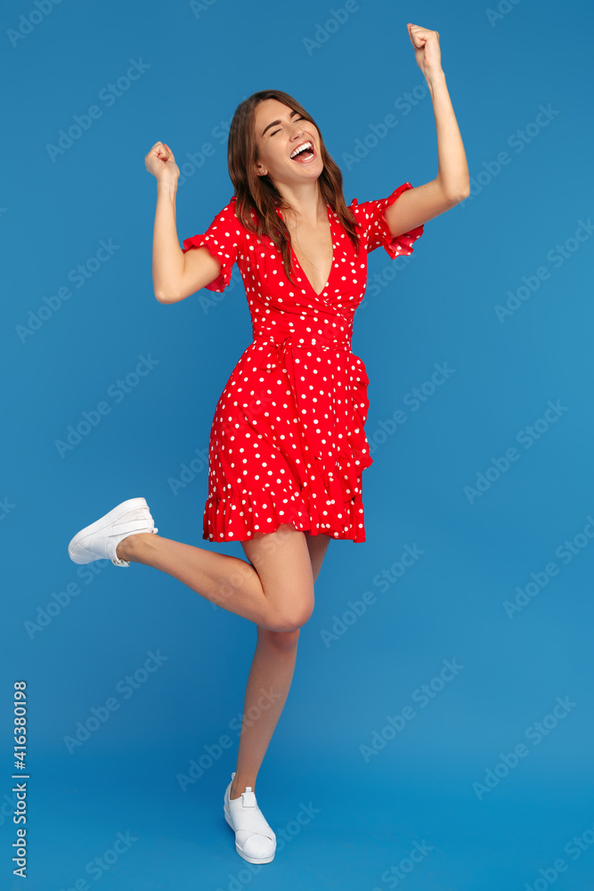 Naklejka premium Successful young woman in red casual dress looking up with excited face with hands up over blue background.