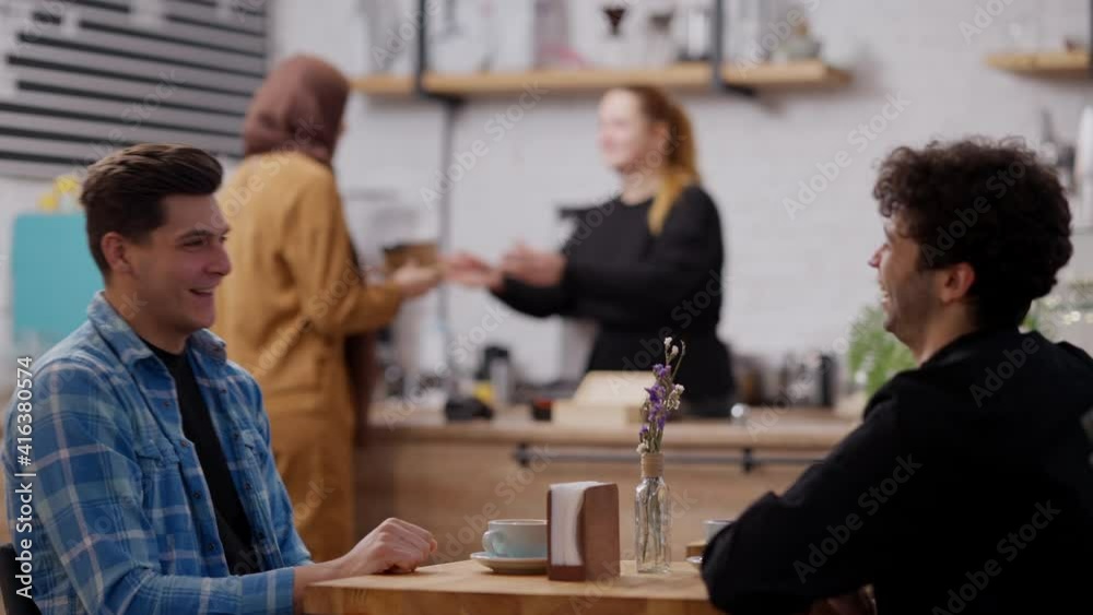 Positive laughing Caucasian and Middle Eastern men talking sitting at table in cafe with blurred worker serving client at background. Happy carefree friends resting in cafeteria. Lifestyle and leisure